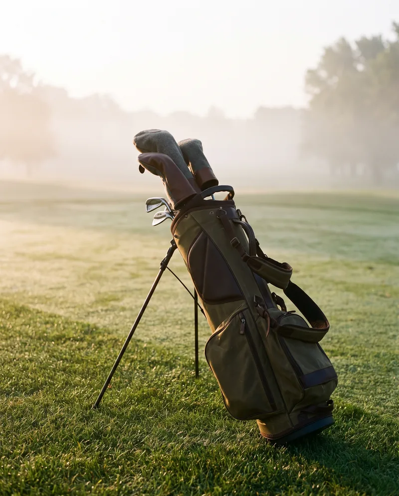 A canvas golf bag on its stand at the first tee, mist across the fairway.